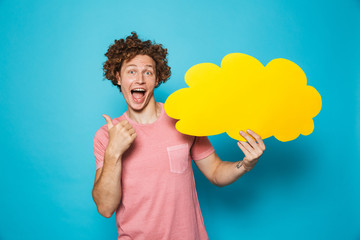 Photo of optimistic curly man 20s holding and pointing finger at yellow copyspace bubble, isolated over blue background