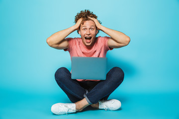 Fototapeta premium Image of hipster man 20s with brown curly hair grabbing head, while sitting on floor with legs crossed, isolated over blue background