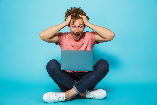 Image of amazed or suprised hipster man 20s with brown curly hair grabbing head, while sitting on floor with legs crossed, isolated over blue background
