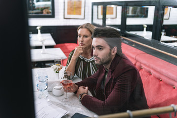 Age difference. Anxious pensive woman touching face and man carrying glass of wine