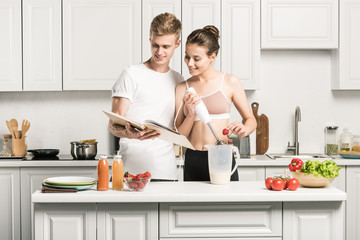 young couple preparing smoothies with recipe book in kitchen