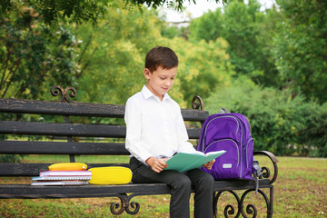 Cute little school child with stationery reading book on bench in park