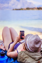 Junge Frau liegt am Strand, Smartphone in der Hand, blaues Wasser im Hintergrund