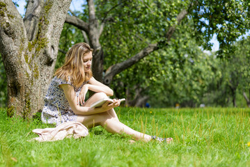 Naklejka na ściany i meble Young cute woman reading a book, sitting in a garden under a tree