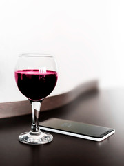 One glass of red wine and a mobile phone on brown wodden table and white blurred wall background. Alcohol drink and smartphone on desk in room.