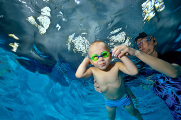 Mother-coach teaches a little boy to swim underwater in the pool and supports him with his hands. The kid looks at the camera and smiles. Portrait. Shooting underwater. Bottom view © alexbard