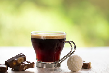 a cup of coffee with chocolate on white wooden table with green leaves background