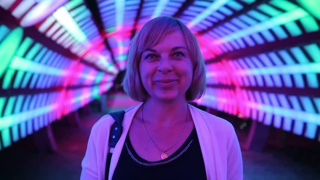 An optimistic view of a happy woman smiling and standing in a half spherical tunnel illuminated with blue, red and white lamps in some club