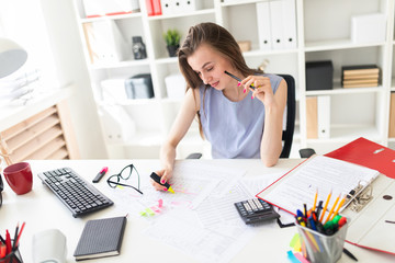 Beautiful young girl in the office sits at a table, holds in her hand a yellow marker and a pencil.