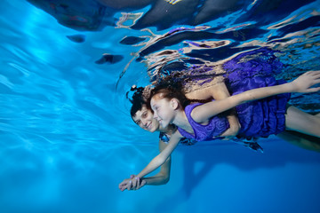 Mother and daughter in purple dress swim forward and hug underwater in the pool on a blue background. Portrait. Side view. Underwater photography. The horizontal orientation of the image