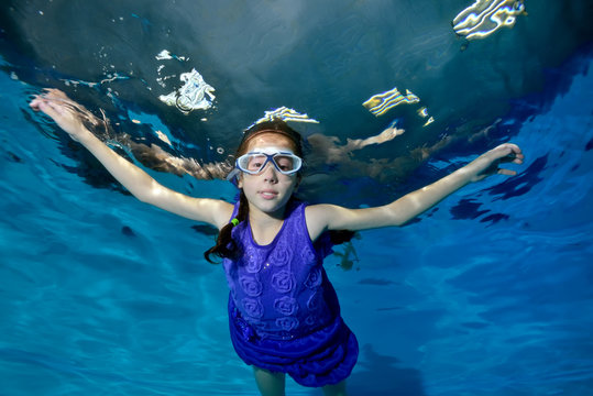 A little girl with pigtails engaged in sports, swims underwater in the pool in a purple swimsuit and looks at the camera. Portrait. Bottom view. The horizontal orientation of the image
