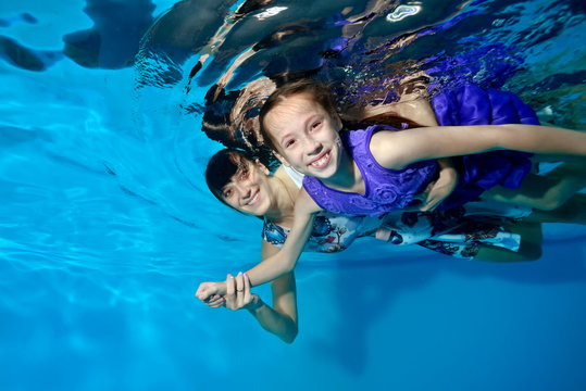 Mom and daughter are laughing, swimming and playing underwater in the pool in beautiful dresses on blue background. Mother hugs daughter. They look at the camera. Portrait. Underwater photography