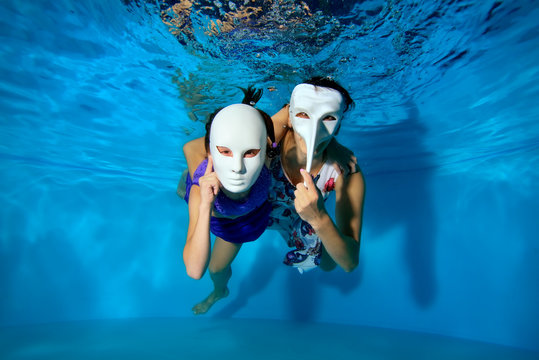 A child and a mother in white masquerade masks swim and pose under the water in the pool in beautiful dresses and look at the camera. Theatrical performance. The horizontal orientation of the image