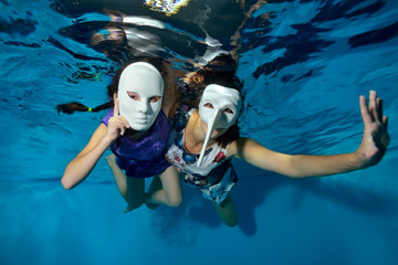 Underwater theater. Two unusual girls swim and play underwater in the pool on a blue background in white masquerade masks in beautiful dresses and look at the camera. Horizontal orientation