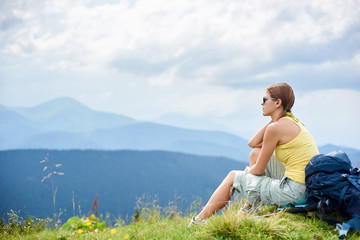 Naklejka premium Back view of attractive woman tourist sitting and resting on grassy hill with backpack. Female backpacker enjoying summer cloudy day in the Carpathian mountains. Outdoor activity, tourism concept