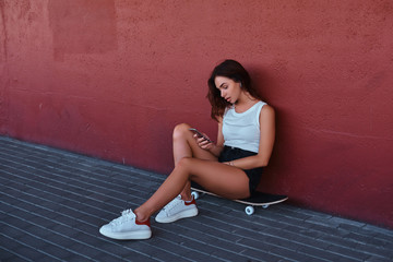 Portrait of a sensual hipster sitting on a skateboard using a smartphone and listening to music while leaning on the wall.