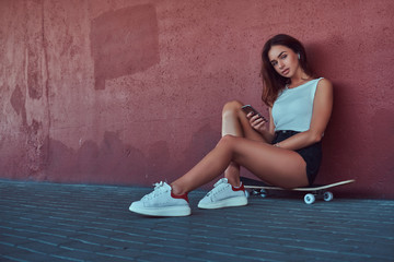 Portrait of a sensual hipster girl dressed in shorts and t-shirt sitting on skateboard holds a...