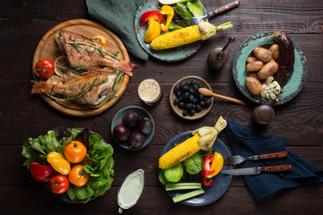 Dinner table with fried fish, corn, salad, grilled potatoes and eggplant, paprika, olives, tomato, celery and sauce, beer top view.
