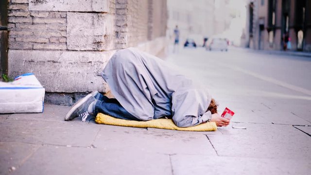 A homeless man begging begs for alms standing on his knees and lower his head to the ground, holding a glass of pennies in his hands in the Streets of Rome, Italy. Homelessness people. 4K (UHD).