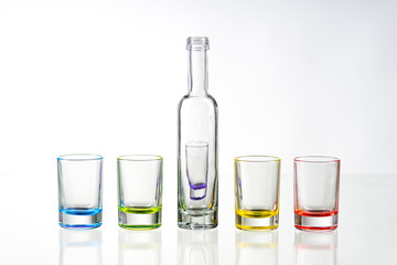 Five multicolored empty shot glasses and small bottle placed symmetrically on a white background.