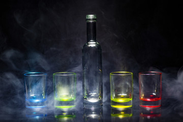 Five multicolored empty shot glasses and small bottle placed symmetrically on a black background in...