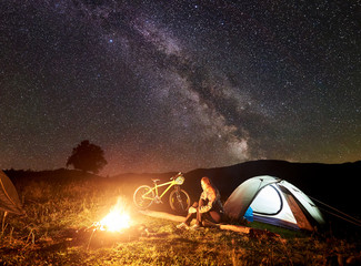 Young woman cyclist having a rest at night camping near burning campfire, illuminated tourist tent, mountain bicycle under amazing beautiful evening sky full of stars and Milky way. Astrophotography
