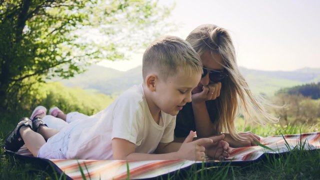 Happy Mother and son playing smartphone under sun light. Green field in park at sunny day, mountain background.
