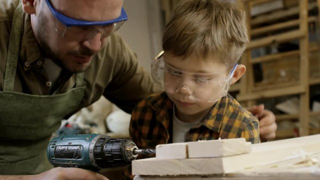 Medium shot of artisan in protective goggle showing son how to use drill in carpentry workshop