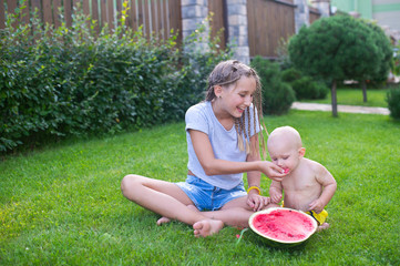 Funny teenage girl feeding her little baby brother with juicy water melon at sunny summer day in green back yard