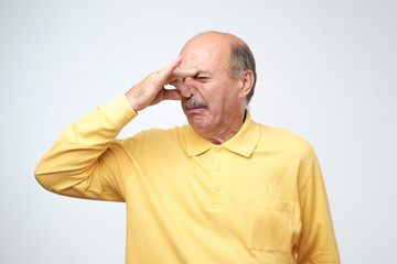 Mature caucasian man in yellow tshirtwith disgust on his face pinches nose.