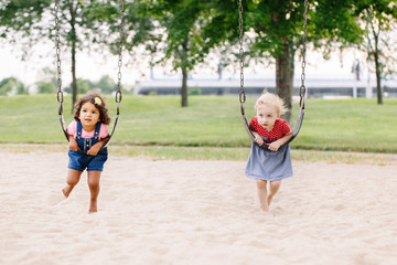 Portrait of two happy smiling little toddlers girls friends swinging on swings at playground...