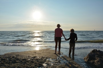 Zwei Kinder stehen Hände haltend  beim Sonnenuntergang am Strand und schauen zum Meer