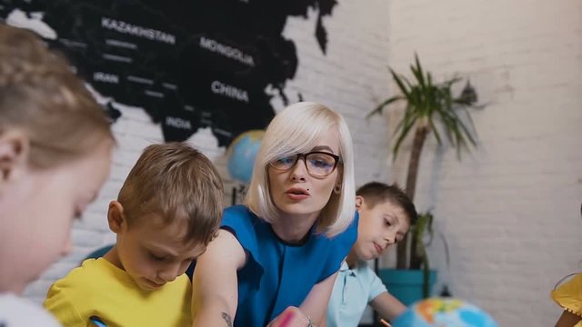 Primary School Or Kindergarten Children Drawing In The Classroom With Teacher Helping. Portrait Of A Young Caucasian Teacher Who Teaches Drawing Their Pupils During Classes Drawing