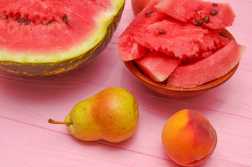 Pieces of watermelon in a plate and a watermelon, a pear, a peach on a pink wooden table