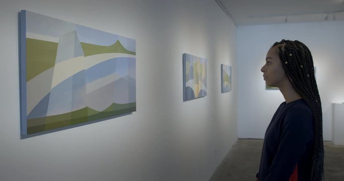 MS Hispanic woman walks into view and looks at abstract landscape paintings in art gallery. Locked off shot, profile view