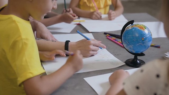 A group of children and a teacher draw on white paper using colored pencils. Primary school children drawing in the classroom with teacher helping