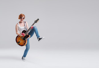 Naklejka premium Young woman musician with an acoustic guitar in hand on a gray background. He laughs and plays rock and roll loudly. Full-length portrait. On the right there is space for text