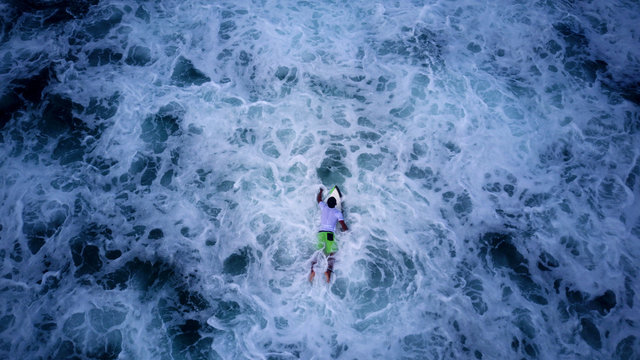 AERIAL SHOT: Surfer paddling on a white board, waiting for a big wave to ride