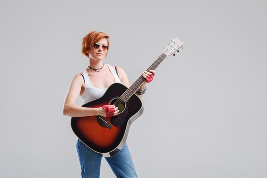 Young woman musician with an acoustic   guitar in hand  on a gray background. He laughs and plays rock and roll loudly. 