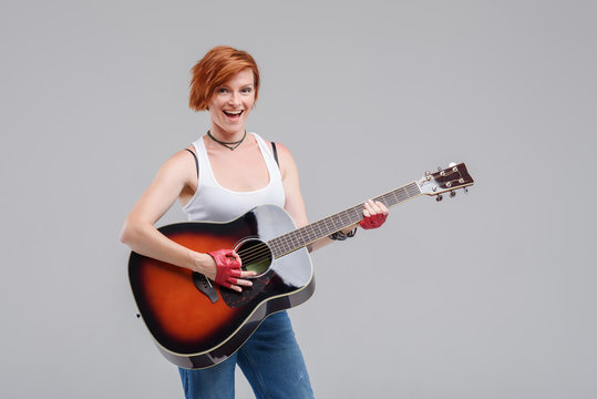 Young woman musician with an acoustic   guitar in hand  on a gray background. He laughs and plays rock and roll loudly. 