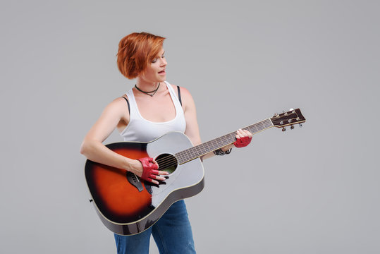 Young woman musician with an acoustic   guitar in hand  on a gray background. He laughs and plays rock and roll loudly. 