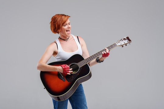 Young woman musician with an acoustic   guitar in hand  on a gray background. He laughs and plays rock and roll loudly. 