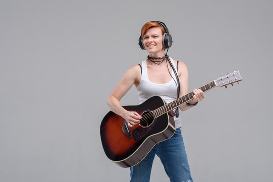 Young woman musician with an acoustic   guitar in hand and in headphones on a gray background. He laughs and plays rock and roll loudly. 