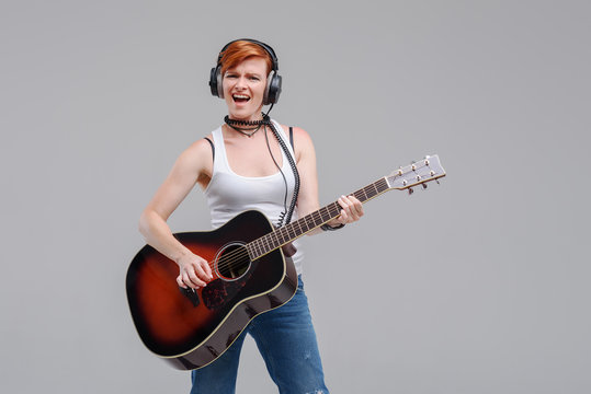 Young woman musician with an acoustic   guitar in hand and in headphones on a gray background. He laughs and plays rock and roll loudly. 