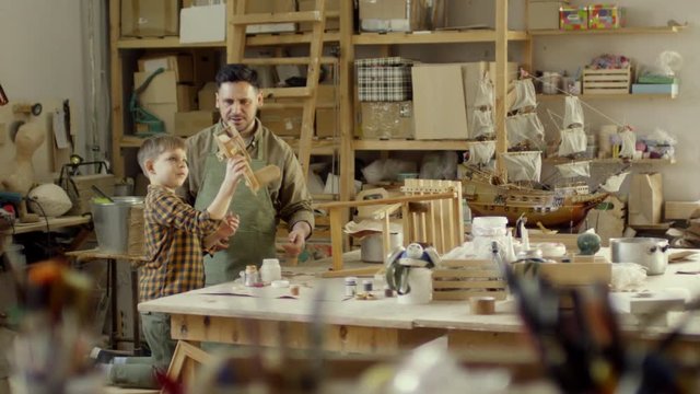 Wide shot with PAN of happy father watching little son play with handmade wooden airplane toy while spending time with him in carpentry workshop