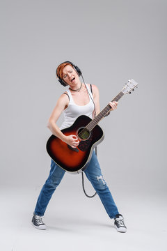 Young woman musician with an acoustic   guitar in hand and in headphones on a gray background. He laughs and plays rock and roll loudly. Full-length portrait