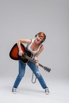 Young woman musician with an acoustic   guitar in hand and in headphones on a gray background. He laughs and plays rock and roll loudly. Full-length portrait