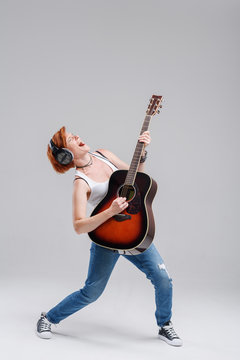 Young woman musician with an acoustic   guitar in hand and in headphones on a gray background. He laughs and plays rock and roll loudly. Full-length portrait