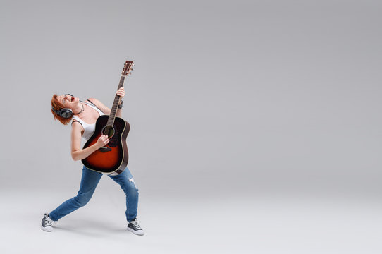 Young woman musician with an acoustic   guitar in hand and in headphones on a gray background. He laughs and plays rock and roll loudly. Full-length portrait. On the right there is space for text