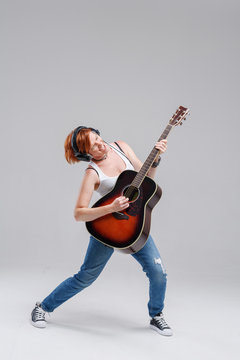 Young woman musician with an acoustic   guitar in hand and in headphones on a gray background. He laughs and plays rock and roll loudly. Full-length portrait